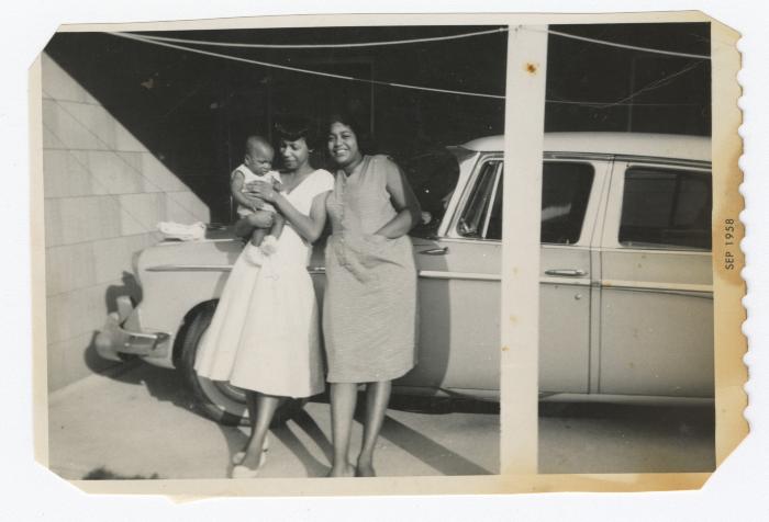 Two women and a baby leaning against a car