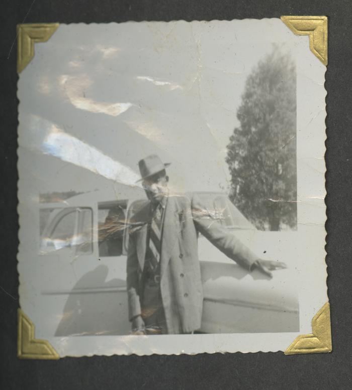 A man in a suit and hat leaning on a car